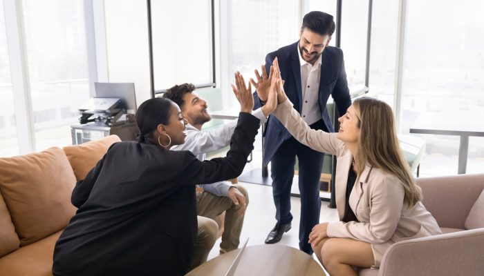Four multinational teammates gathered together in modern coworking space giving high five, gesture of unity and teamwork, celebrating joint achievement, closing deal, completing project successfully